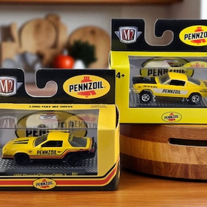May include: Two die-cast model cars in yellow and black Pennzoil packaging. The cars are 1985 Chevrolet Camaro IROC-Z models. The packaging includes the Pennzoil logo and the text "LONG MAY WE DRIVE".
