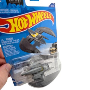 K&ouml;nnte beinhalten: Ein Hot Wheels Spielzeugauto aus Druckguss in einer transparenten Kunststoffverpackung. Das Auto ist ein graues Batwing-Fahrzeug mit schwarzen Fl&uuml;geln und gelben Akzenten. Die Verpackung zeigt das Hot Wheels-Logo und das Batman-Branding.