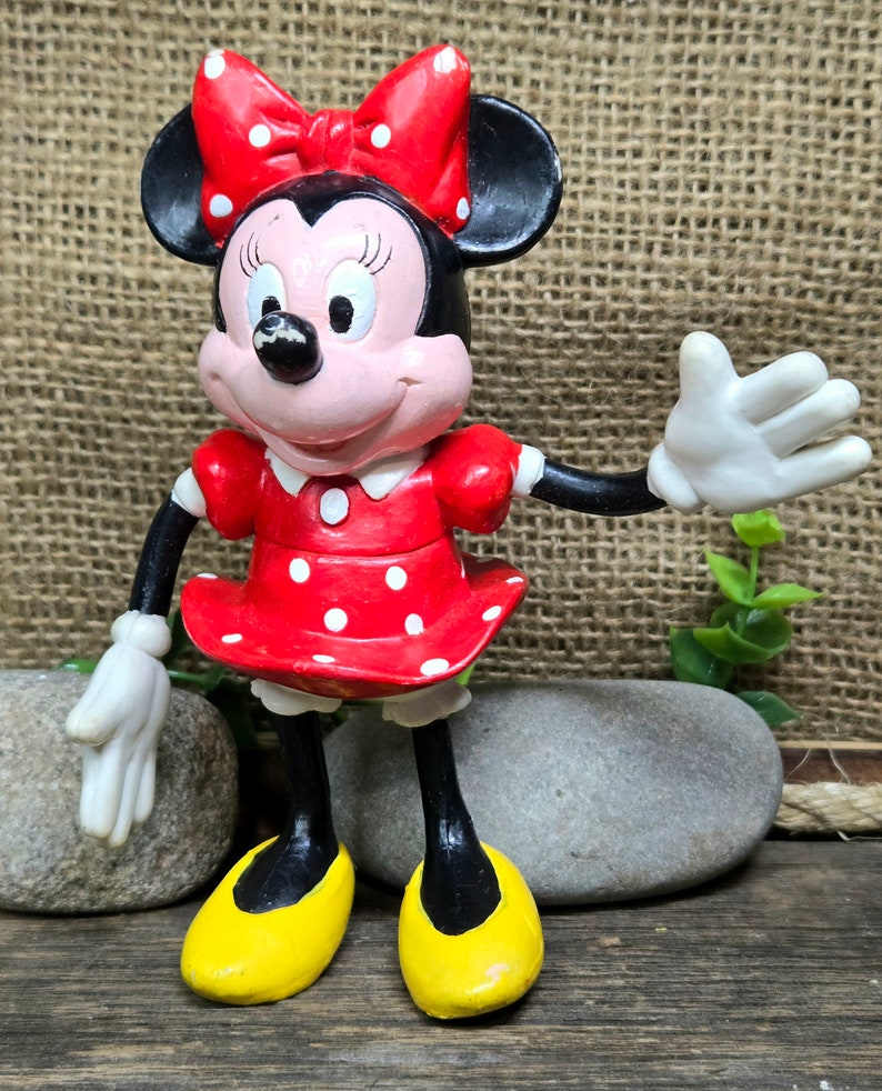 May include: A red and white polka dot dress with a black bow and white gloves. The figure is standing on a wooden surface with two gray rocks.