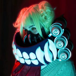 May include: A cosplay ensemble featuring a character with light green hair and eyes. The costume includes a mask, a collar with large white teeth, and a neck piece with glowing blue accents. The outfit is completed with a red scarf and a green sweater.