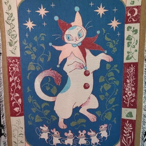May include: A woven throw blanket with a whimsical design. The central image features a cat-like figure in a jester's outfit, with stars and floral elements on a blue background. The border has a brown fringe.