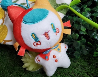 Puede incluir: Un peluche con un diseño caprichoso. El juguete presenta una cabeza roja y amarilla con una cara blanca, detalles turquesa y detalles bordados. Un pequeño peluche blanco en forma de estrella está adjunto debajo. El juguete está sobre un fondo verde.