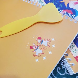 以下が含まれることがあります： 黄色いプラスチック製のスクレーパーが、スパイラルバインディングの付いた黄色のノートの上に置かれています。ノートには、星と「LENLIPUI」という文字と共に、踊る2匹の白いネズミのデザインが施されています。