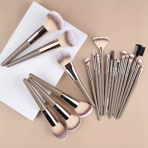 Op de afbeelding: Een set make-upkwasten met lichtbruine handvatten en metalen hulzen. De set bevat diverse vormen kwasten, waaronder grote poederkwasten, schuine kwasten, een waaierkwast, een spoolie en een wimperkam met zachte synthetische haren.