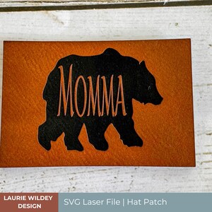 May include: A brown leather patch with a black bear silhouette and the word "MOMMA" inside the bear. The patch is for a hat or other apparel.