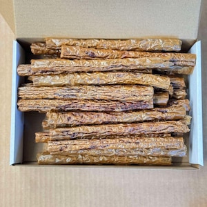 May include: A box filled with long, thin, brown dog chews. The chews are made of dried meat and are stacked neatly in the box.