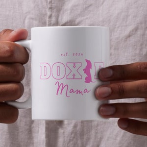 Person holding a white Doxie Mama mug with pink lettering, against a light background.