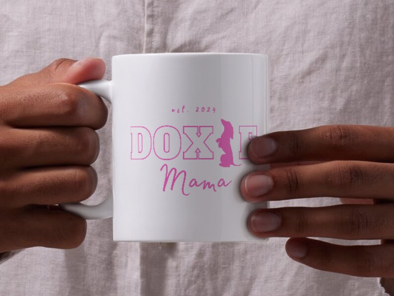 Person holding a white Doxie Mama mug with pink lettering, against a light background.