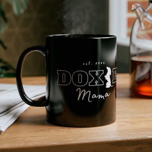 Black Doxie Mama mug with white lettering on a wooden table next to a newspaper and a pot of tea.