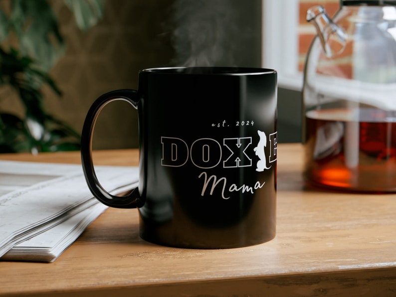 Black Doxie Mama mug with white lettering on a wooden table next to a newspaper and a pot of tea.