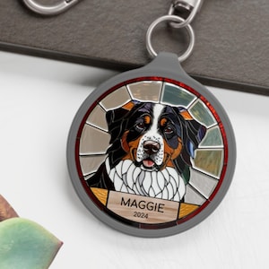 May include: A round, gray keychain featuring a stained-glass style image of a Bernese Mountain Dog. The dog has black, brown, and white markings. The name "MAGGIE" and the year "2024" are printed on a wooden-look banner.