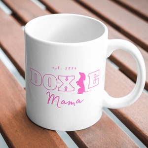 White Doxie Mama mug with pink lettering on a wooden outdoor table.