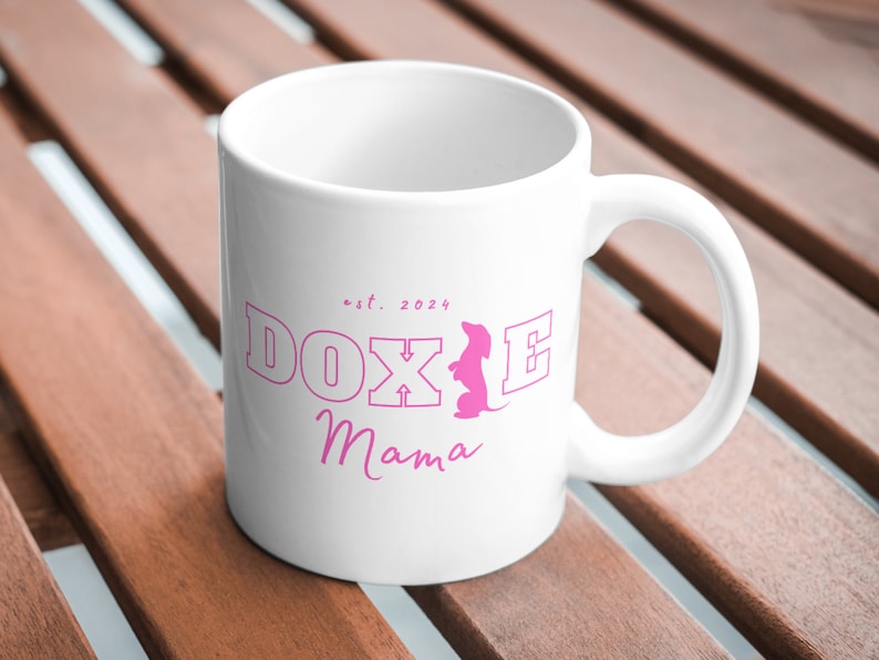 White Doxie Mama mug with pink lettering on a wooden outdoor table.