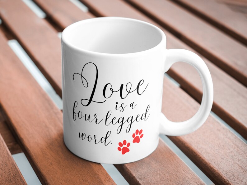 May include: White ceramic coffee mug with black text that reads "Love is a four legged word." Two red paw prints are below the text.