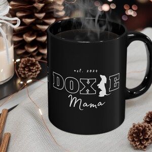 Black Doxie Mama mug with white lettering in a cozy setting with pinecones and a candle.