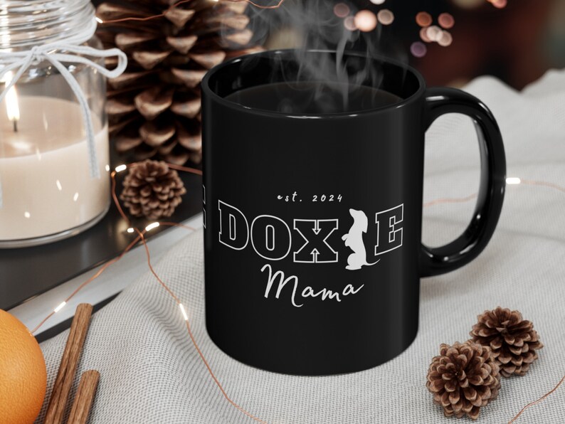 Black Doxie Mama mug with white lettering in a cozy setting with pinecones and a candle.