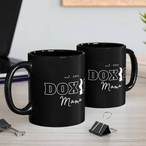 Two black Doxie Mama mugs with white lettering on a modern office desk, surrounded by office supplies.