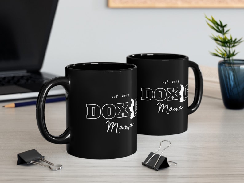Two black Doxie Mama mugs with white lettering on a modern office desk, surrounded by office supplies.