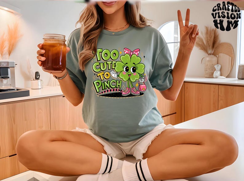 以下が含まれることがあります： 青いTシャツを着た女性。Tシャツには緑色の四つ葉のクローバーのグラフィックと「Too Cute To Pinch」の文字がプリントされています。白いショートパンツと白黒のストライプの靴下を履いています。