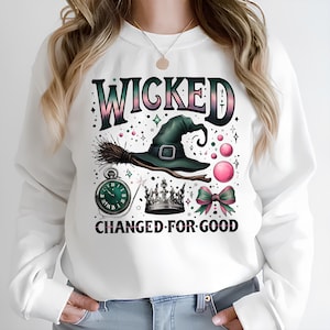 Könnte beinhalten: Weißer Pullover mit einem grünen Hexenhut, einem Besen, einer Krone, einer Schleife und einer Taschenuhr. Der Text "WICKED CHANGED FOR GOOD" ist auf der Vorderseite aufgedruckt.