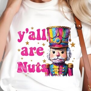 Peut inclure: T-shirt blanc avec une illustration colorée d'un soldat casse-noisette et le texte rose "y'all are Nuts". Le casse-noisette a un visage détaillé, un grand chapeau et un uniforme. Des étoiles dorées sont dispersées autour du texte.