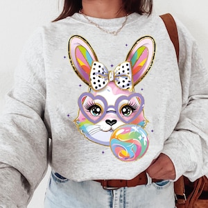 Puede incluir: Una sudadera gris con un conejito de dibujos animados colorido que lleva gafas en forma de corazón y un lazo de lunares. El conejito sostiene una pelota de colores arcoíris.