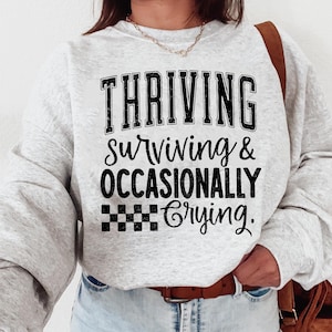 May include: A gray sweatshirt with black text that reads "THRIVING surviving & OCCASIONALLY crying."  The text is in a vintage style font.  The sweatshirt has a checkered pattern below the text.