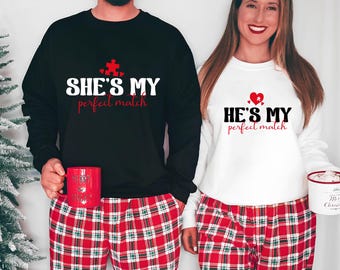 May include: A couple wearing matching black and white sweatshirts with red text. The text on the black sweatshirt says "She's my perfect match" with a red puzzle piece. The text on the white sweatshirt says "He's my perfect match" with a red heart. Both are wearing red and black plaid pajamas.