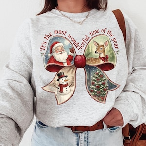 Peut inclure: Un sweat-shirt gris clair avec un motif de nœud de Noël. Le nœud comprend des illustrations du Père Noël, d'un cerf, d'un bonhomme de neige et d'un sapin de Noël. Le texte "It's the most wonderful time of the year" est imprimé sur le nœud.