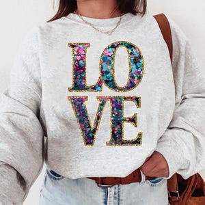 Peut inclure: Un sweat-shirt gris avec le mot "LOVE" écrit en grandes lettres scintillantes. Les lettres sont faites de paillettes irisées dans les tons rose, bleu et vert.