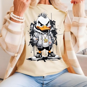 Peut inclure: Un t-shirt crème avec une illustration de canard en dessin animé portant un sweat-shirt gris, une chaîne en or et un pendentif en or avec la lettre 'D'. Le canard est debout sur un fond noir et blanc avec des éclaboussures de peinture.