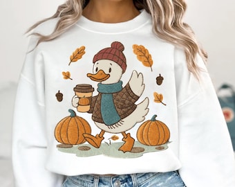 Op de afbeelding: Witte sweatshirt met een cartoon eend met een hoed, sjaal en jas, met een koffiekopje met een pompoenontwerp. Het ontwerp bevat pompoenen, bladeren en eikels, wat een herfstthema oproept.