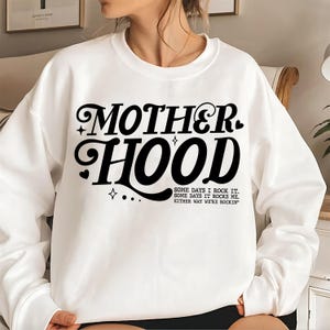 May include: White sweatshirt with black text that reads "MOTHERHOOD" in a retro font. The text is followed by the phrase "SOME DAYS I ROCK IT. SOME DAYS IT ROCKS ME. EITHER WAY WE'RE ROCKIN'"