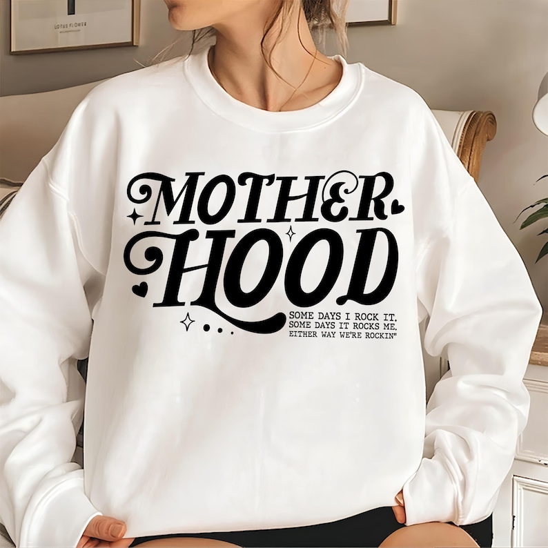 May include: White sweatshirt with black text that reads "MOTHERHOOD" in a retro font. The text is followed by the phrase "SOME DAYS I ROCK IT. SOME DAYS IT ROCKS ME. EITHER WAY WE'RE ROCKIN'"
