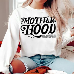 May include: White sweatshirt with black text that reads "MOTHERHOOD" in a retro font. The text is followed by the phrase "SOME DAYS I ROCK IT. SOME DAYS IT ROCKS ME. EITHER WAY WE'RE ROCKIN'"