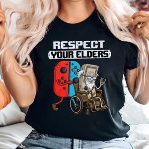 May include: Black t-shirt with the text "RESPECT YOUR ELDERS". The graphic shows a red and blue game controller pushing a vintage video game character in a wheelchair. The character is holding a small device.