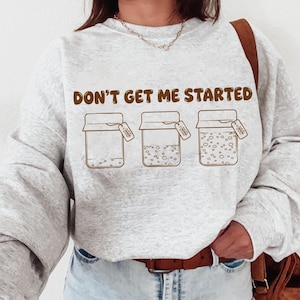 May include: A light gray sweatshirt with the phrase "DON'T GET ME STARTED" in brown lettering. Below the text are three jars with varying contents and tags. The sweatshirt is paired with blue jeans and a brown leather bag.