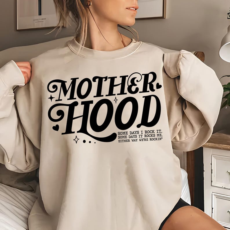 May include: A beige sweatshirt with the text "MOTHERHOOD" in black stylized lettering. The text below reads "SOME DAYS I ROCK IT. SOME DAYS IT ROCKS ME. EITHER WAY WE'RE ROCKIN'"