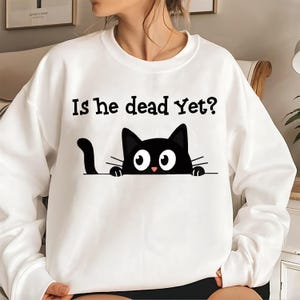 Peut inclure: Sweat-shirt blanc avec l'inscription "Is he dead Yet ?" et un chat noir de dessin animé. Le chat a de grands yeux blancs, un nez rose et une queue noire, regardant par-dessus une ligne.