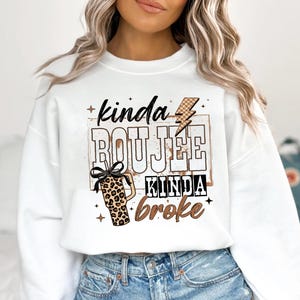 May include: White sweatshirt with the text "kinda BOUJEE kinda BROKE" in a brown and black design. The design includes a lightning bolt, a leopard print tumbler with a black bow, and stars.