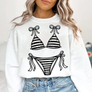 Peut inclure: Sweat-shirt blanc avec un motif de bikini à rayures noires et blanches. Le haut du bikini a des nœuds sur les bretelles, et le bas a des nœuds sur les côtés. Un design estival et amusant.