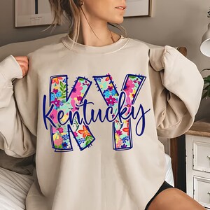 May include: A beige sweatshirt with a floral design of the letters "KY" and the word "Kentucky" in blue.