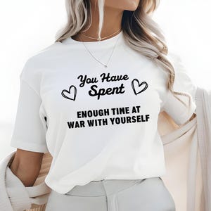May include: White t-shirt with black text that reads "You Have Spent Enough Time At War With Yourself". The text is surrounded by two small hearts.