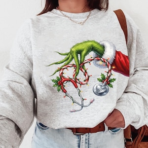 Peut inclure: Un sweat-shirt gris clair avec un motif sur le thème du Grinch. Le motif comprend une main verte, un stéthoscope rouge et blanc avec du houx et des lumières, et un bonnet de Père Noël. Le sweat-shirt est en matière douce.