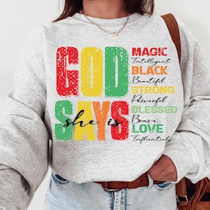 Peut inclure: Un sweat-shirt gris avec les mots "God Says She Is" en lettres majuscules colorées. Les mots "Magic, Intelligent, Black, Beautiful, Strong, Powerful, Blessed, Brave, Love, Influential" sont listés verticalement en lettres plus petites.