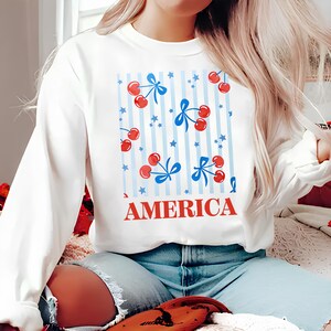 Peut inclure: Sweat-shirt blanc avec un motif rayé bleu et blanc, des cerises rouges, des nœuds bleus et des étoiles blanches. Le texte "AMERICA" est imprimé en lettres rouges sur le devant du sweat-shirt.