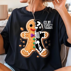 Può includere: T-shirt nero con un disegno di omino di pan di zenzero diviso per rivelare uno scheletro. L'omino di pan di zenzero ha un papillon viola e bottoni verdi. Il testo "CUT MY LIFE INTO PIECES" è stampato sulla maglietta.