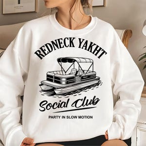 Peut inclure: Sweat-shirt blanc avec l'inscription "REDNECK YAKHT Social Club" au-dessus d'une illustration de bateau. L'expression "PARTY IN SLOW MOTION" est imprimée sous le dessin du bateau. Le sweat-shirt est de style décontracté et confortable.