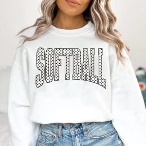 Peut inclure: Sweat-shirt blanc avec le mot "SOFTBALL" en grandes lettres noires à pois sur le devant. Le sweat-shirt a un col rond et des manches longues. Le modèle porte un jean bleu.