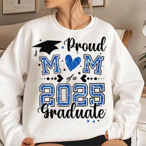 May include: White sweatshirt featuring the text "Proud Mom of a 2025 Graduate" in black script and blue block letters. The design includes a graduation cap, a blue heart, and stars. A casual top for graduation celebrations.